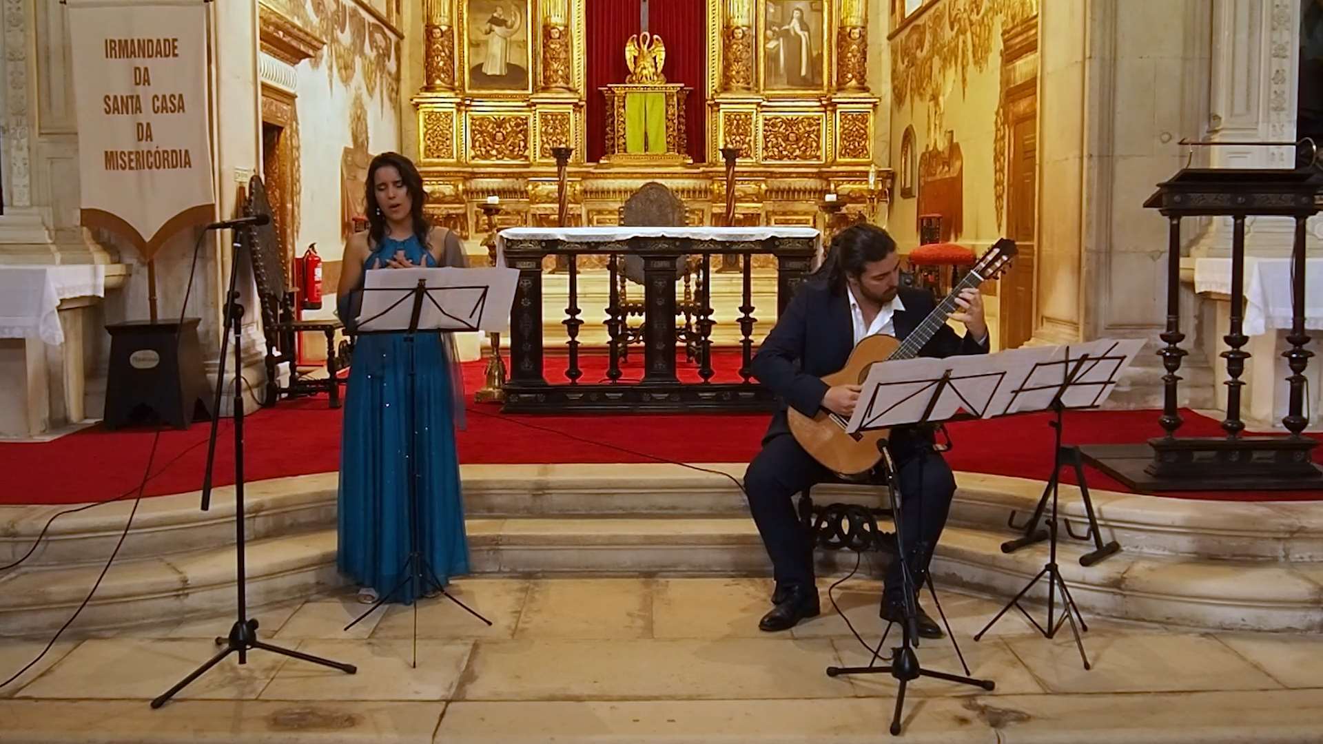 Numa igreja antiga, estão um rapaz e uma rapariga. A rapariga está de pé, à esquerda a cantar, e o rapaz está à direita sentado a tocar guitarra clássica. Os dois estão com roupa formal.